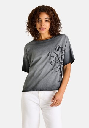 Frau mit lockigem Haar, die ein gewaschenes schwarzes Kurzarmshirt mit einem schwarzen bestickten Blattmuster und weißen Hosen trägt.