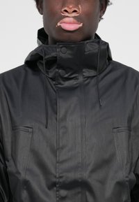 Veste noire légère avec un col montant, capuche avec cordon de serrage, deux poches avant et fermetures à boutons-pression. Texture lisse et mate.