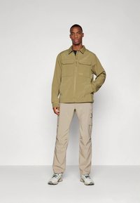 Veste zippée kaki avec deux poches poitrine, associée à un pantalon cargo beige et des baskets grises. Design simple, tissu léger et détails pratiques.