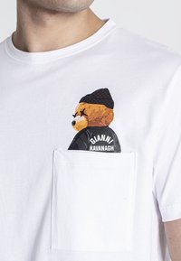 Bílá bavlněná tričko s grafikou medvěda v černé čepici a bundě s nápisem "GIANNI KAVANAGH" na oblasti kapsy.