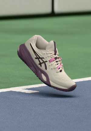 Scarpa sportiva beige e viola con logo nero e dettagli rosa sulle stringhe che fluttua sopra la superficie di un campo da tennis vicino alle linee bianche.