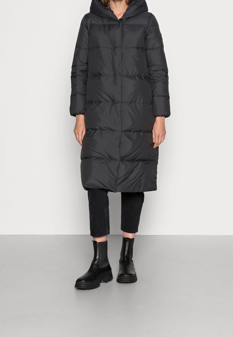 Svart puffjacka med huva, med en quiltad design och långa ärmar, i kombination med mörka cropped byxor och kraftiga svarta ankelboots.
