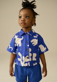 Camicia azzurra a maniche corte con motivi animali bianchi e tasca, abbinata a pantaloni corti blu coordinati. Il tessuto ha una finitura testurizzata.