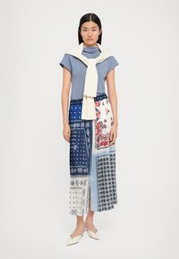 T-shirt blu con collo a imbuto, maglione crema legato sulle spalle e gonna maxi a patchwork con motivi blu, bianco e rosso. Scarpe con tacco bianche.