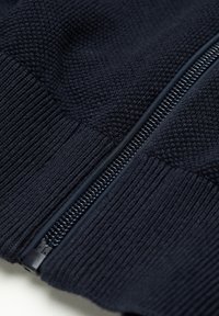 Primo piano di un tessuto di maglia di colore blu scuro con una cerniera in plastica parzialmente chiusa, che mostra motivi a coste e trame di maglia.