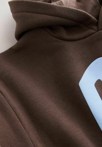 Hnědá mikina s kapucí, která má na přední straně velký světle modrý grafický motiv, vyrobená z měkkého, texturovaného materiálu s elastickým detailem límce.