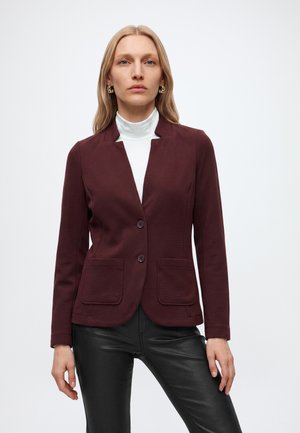 Vrouw die een bordeaux blazer draagt over een witte coltrui en zwarte leren broek, staand tegen een eenvoudige lichte achtergrond.