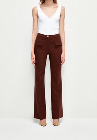 Žena v hnědých kalhotách s vysokým pasem a širokými nohavicemi s předními kapsami, bílé bezrukávové topu s výstřihem do V a béžových botách s špičatou špičkou.