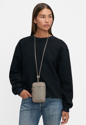 Femme portant un sweat à capuche noir ample et un jean, avec une pochette grise en bandoulière suspendue à une longue sangle autour de son cou.