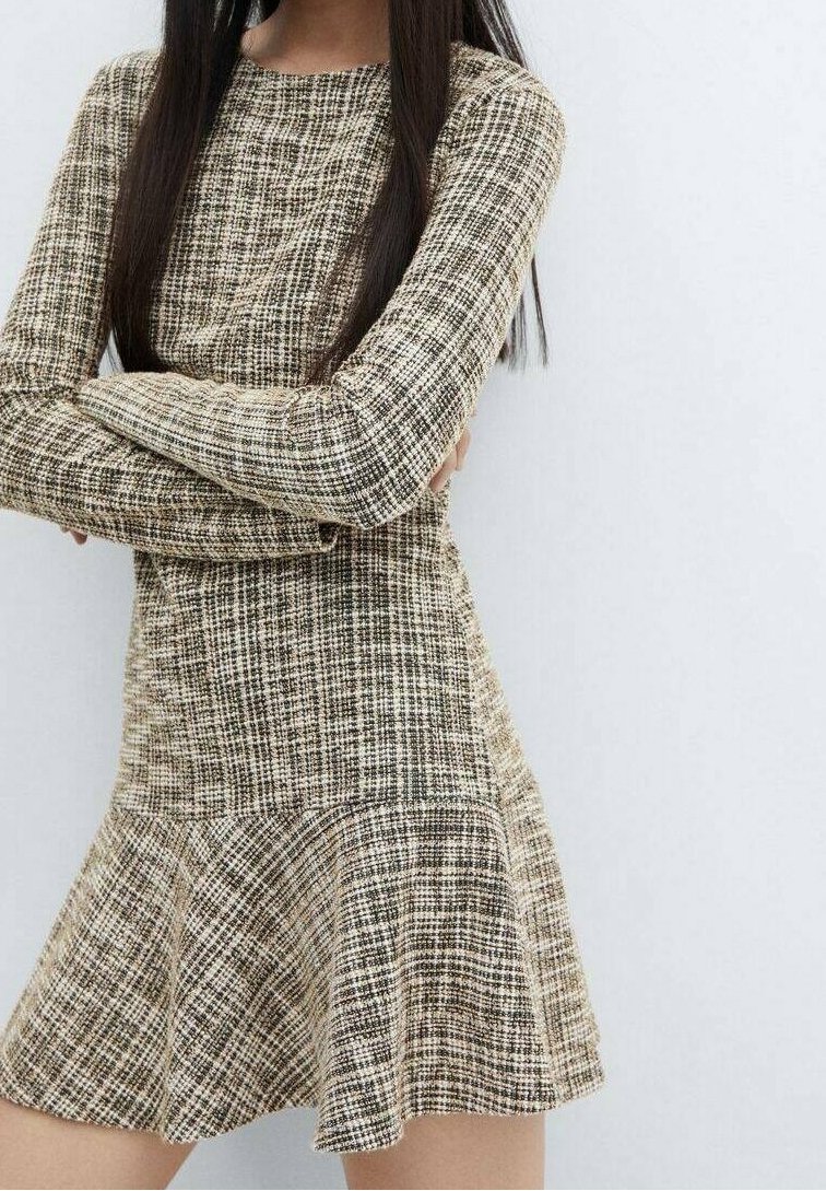 Femme portant une robe à manches longues à carreaux beige et noir avec une jupe évasée, debout devant un fond clair uni.