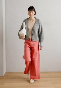 Šedý žebrovaný cardigan s knoflíky, béžový top a růžové široké kalhoty s vysokým pasem. Drží bílou sochařskou kabelku. Krémové boty.