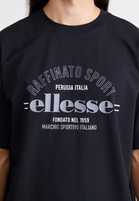 Svart t-skjorte med rund hals, korte ermer og frontgrafikk i hvitt og grått: "Raffinato Sport" og "ellesse."
