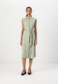Sleeveless dress in light green linen with a button-down front, matching belt at the waist, and knee-length hem. Black heeled shoes.