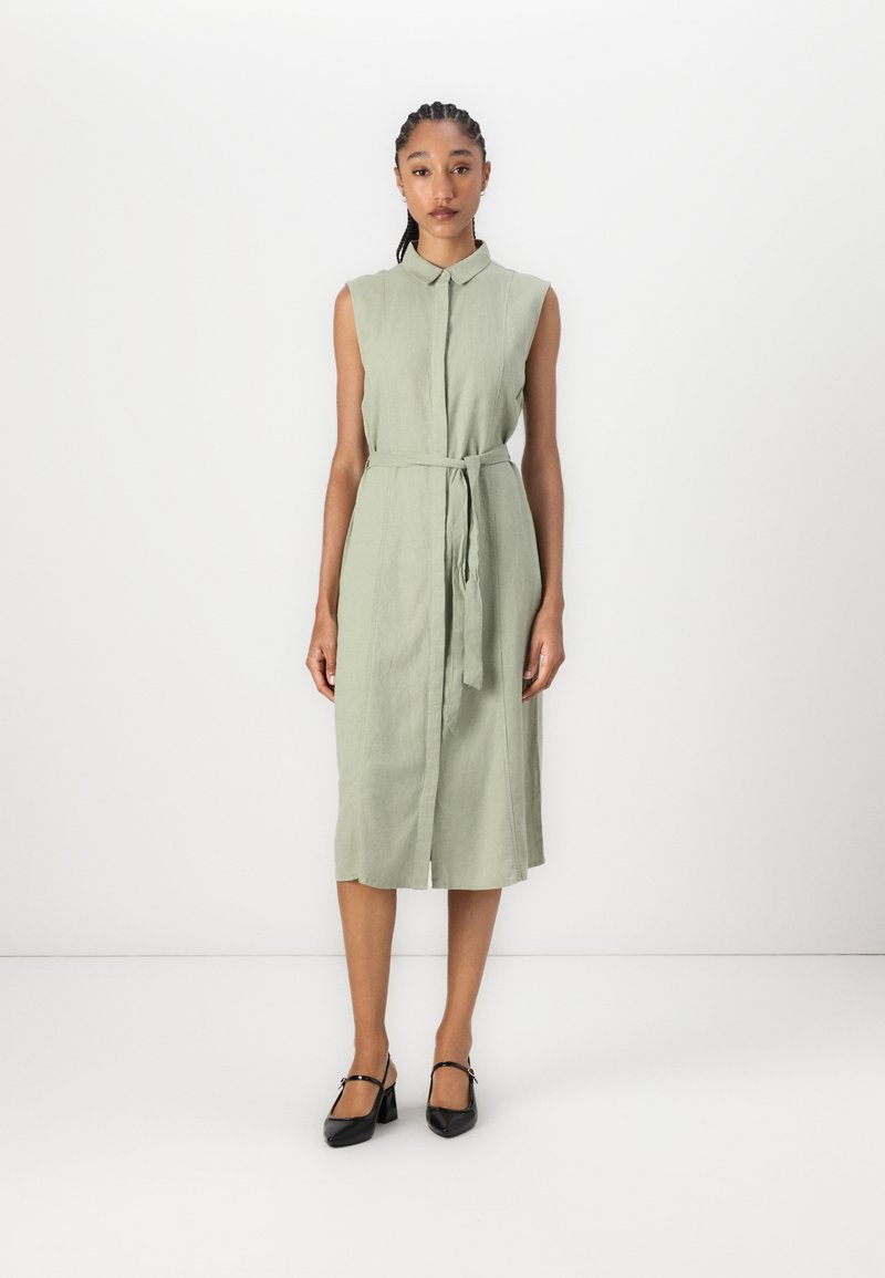 Sleeveless dress in light green linen with a button-down front, matching belt at the waist, and knee-length hem. Black heeled shoes.