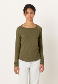 Lokkis juustega naine kannab oliivirohelist pikkade varrukatega pusa ja helerohelisi teksapükse, seistes ühevärvilise valkja tausta ees.