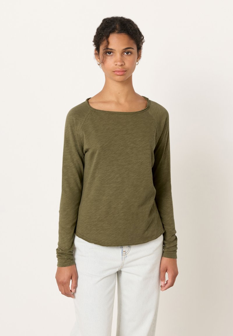 Lokkis juustega naine kannab oliivirohelist pikkade varrukatega pusa ja helerohelisi teksapükse, seistes ühevärvilise valkja tausta ees.