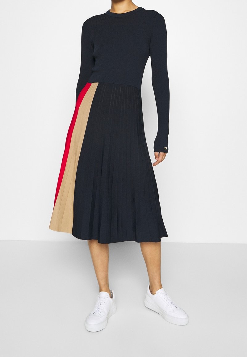 Femme portant un haut noir à manches longues et une jupe midi plissée avec des panneaux noirs, beige et rouges, assortie à des baskets blanches.