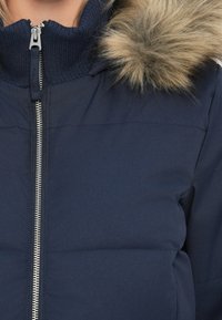 Námornícky modrá péřová bunda s vysokým límcem, se stříbrným zipem a kapucí podšitou kožešinou. Materiál je hladký s mírně lesklou texturou.