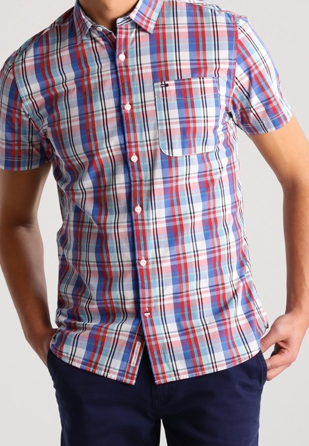 Homme portant une chemise à manches courtes à carreaux rouges, bleus et blancs avec une poche avant, associée à un pantalon bleu foncé.
