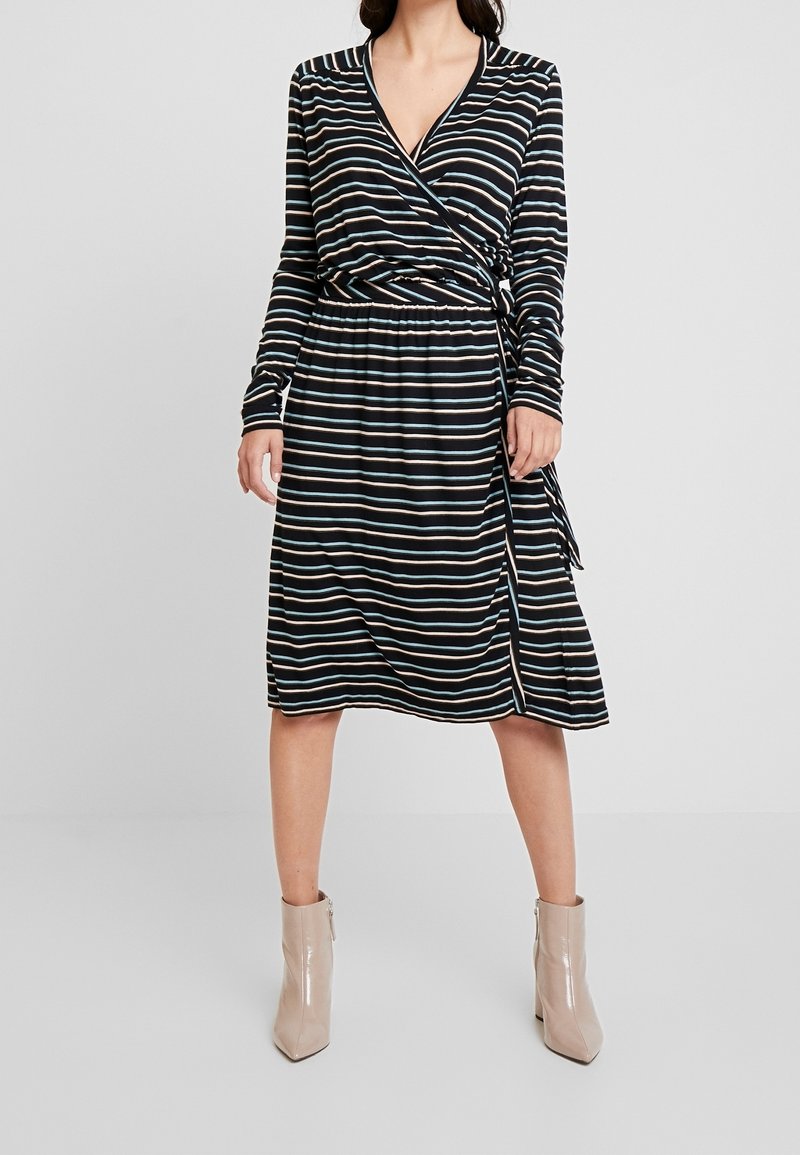 Gestreepte wikkeljurk in zwart, met lange mouwen, een getailleerde taille en een zijsplit; gecombineerd met beige enkellaarzen.