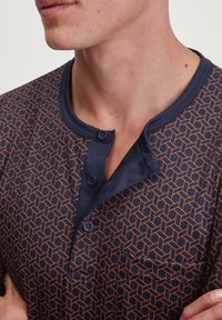 Chemise bleu marine avec un motif géométrique orange, dotée d'un col Henley, de deux boutons et d'une poche sur le côté gauche. Texture de tissu douce.