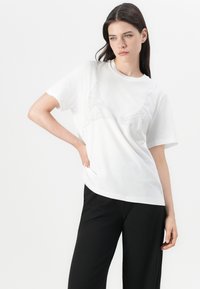 White cotton T-shirt with short sleeves and a scoop neckline, featuring a decorative ribbed design across the chest. Black loose-fitting pants.