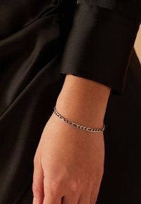 Zilveren kettingarmband om een arm, met schakels van verschillende maten. De achtergrond bestaat uit zacht gedrapeerd zwart stof.