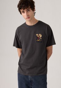 Camiseta de algodón gris con un pequeño diseño bordado de un personaje que lleva un sombrero de ala ancha, combinada con unos jeans de mezclilla azul.