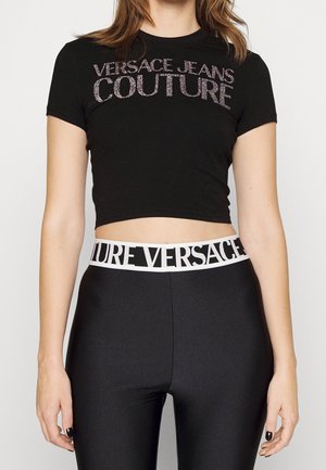 Kobieta ubrana w czarny krótki t-shirt z błyszczącym napisem "Versace Jeans Couture" oraz czarne legginsy z białym paskiem "Versace" w talii.