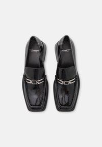 Mocassins en cuir verni noir avec bout carré, accent à boucle argentée et bordure texturée contrastante. Design élégant et moderne.