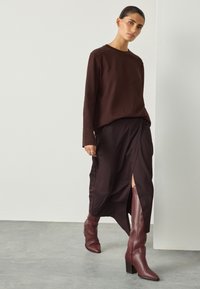 Haut marron à manches longues en tissu texturé, associé à une jupe foncée rayée avec une fente sur le côté. Bottes bordeaux montant jusqu'aux genoux avec un talon carré.