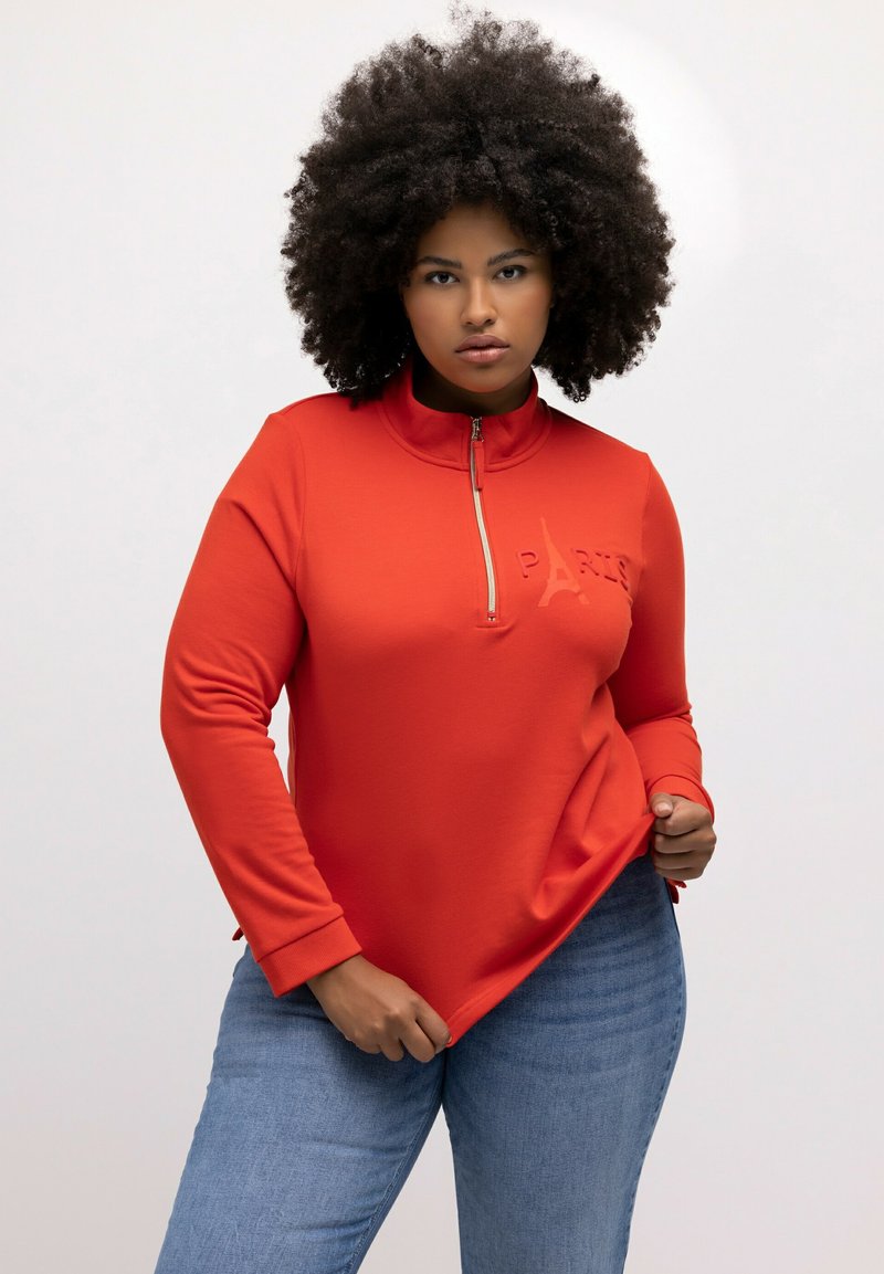 Sudadera roja con cremallera y letras en relieve "PARIS", cuello alto y mangas largas. Combinada con jeans azules de corte relajado.