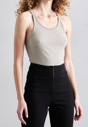 Femme portant un débardeur beige côtelé et un pantalon noir taille haute avec une fermeture éclair à l'avant, debout devant un fond clair uni.