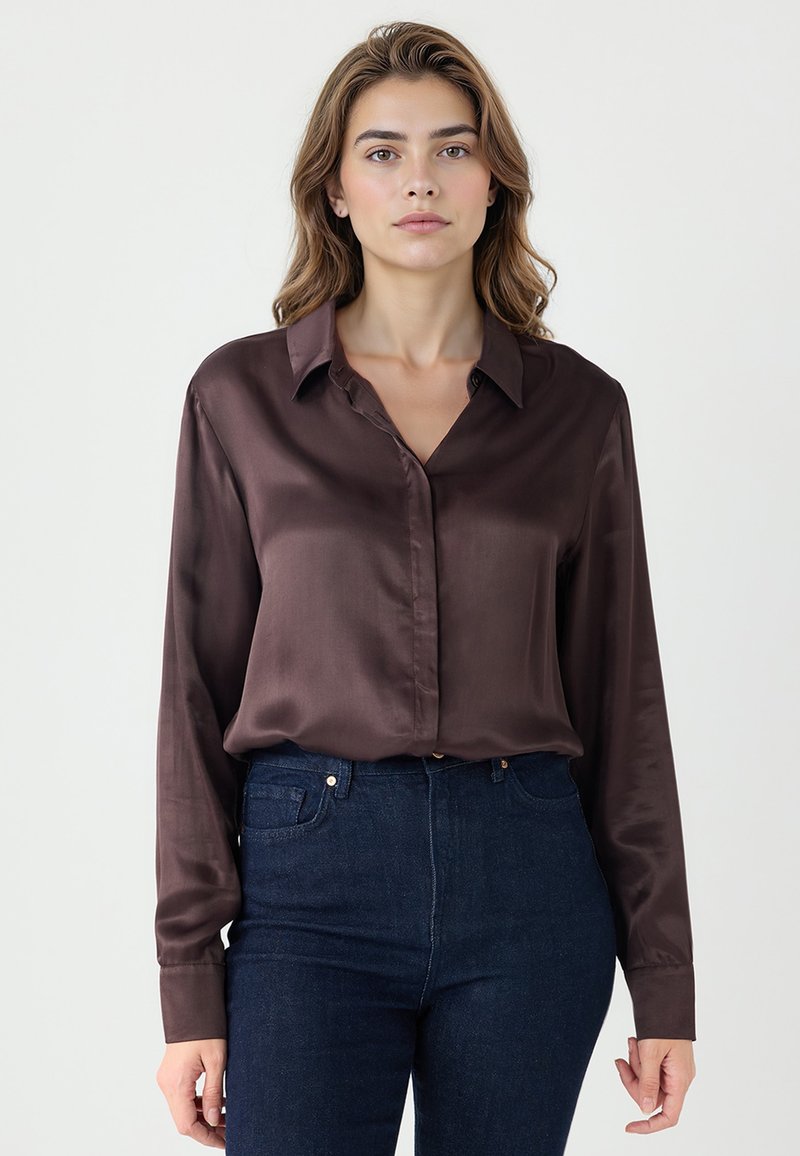 Jeune femme portant un chemisier en satin brun foncé et un jean bleu foncé taille haute, se tenant devant un fond clair uni.