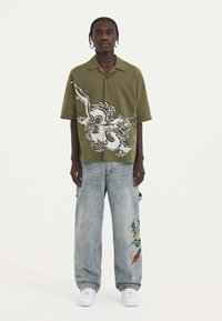 Olivgrön kortärmad skjorta med vit drakegrafik, tillsammans med ljusblå baggyjeans med färgglad broderi på benet. Vita sneakers.