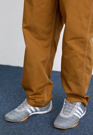 Grijze sportschoenen met witte strepen en een geblokte textuur, gecombineerd met losse bruine pants met witte stiksels.