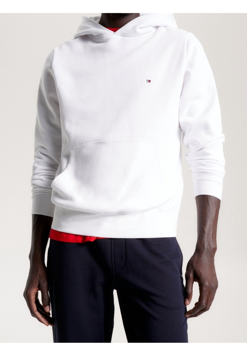 Homme portant un sweat à capuche blanc avec une poche avant, petit logo sur la poitrine, chemise rouge en dessous et pantalon sombre sur un fond clair uni.