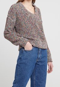 Mehrfarbiger gestrickter Pullover mit V-Ausschnitt, langen Ärmeln und einer groben Textur. Kombiniert mit hoch geschnittenen blauen Jeans.
