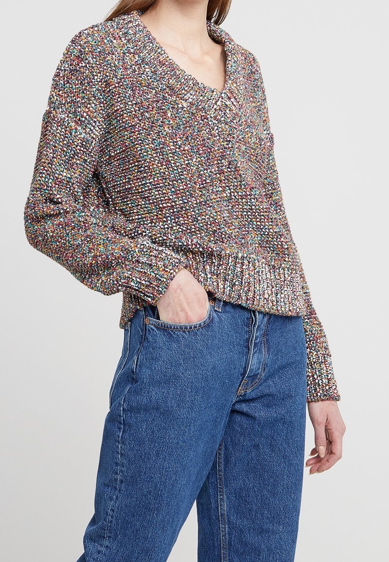 Maglione lavorato a maglia multicolore con scollatura a V, maniche lunghe e una texture spessa. Abbinato a jeans in denim blu a vita alta.