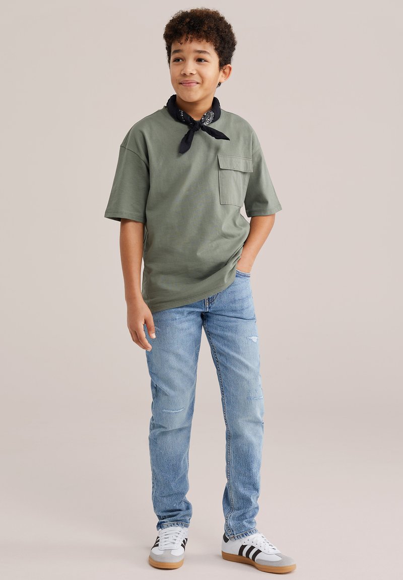 Zēns stāv ar roku kabatā, valkājot zaļu T-kreklu, zilas džinsas, baltus sporta apavus un melnu kaklasaiti uz vienkārša fona.