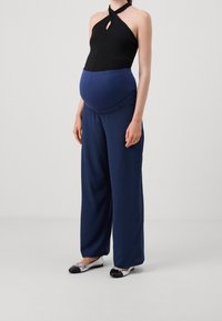 Zwart haltertop met sleutelgatdetail, blauwe paneel en marineblauwe broek met wijde pijpen van textuurstof; zilveren en zwarte platte schoenen.