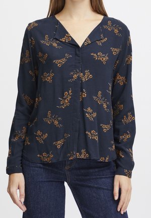 Vrouw draagt een donkerblauwe blouse met een bruin bloemenprint, V-halskraag en lange mouwen, gecombineerd met een donkere blauwe spijkerbroek tegen een witte achtergrond.