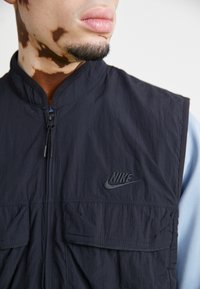 Černá bezrukávová vesta Nike vyrobená z lehké, texturované látky, s předním zipem a dvěma velkými předními kapsami.