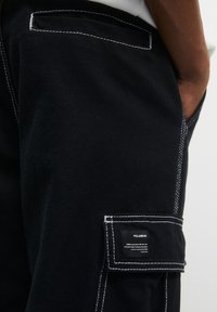 Zwarte denim cargo pants met witte stiksels, zijzakken en een labelpatch. De stof lijkt duurzaam met een klassieke snit.