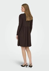 Bruine lange mouw jurk met een aansluitend lijfje en een uitlopende rok. Het stof heeft een geribbeld patroon, aangevuld met zwarte, hoge sling-back schoenen.