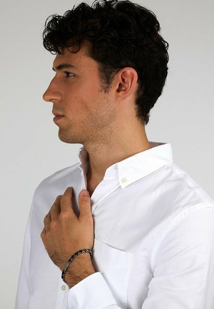 White button-up shirt with a collar, featuring a left chest pocket. Notable black bracelet on the wrist, minimal wrinkles, neutral background.