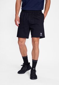 Black athletic shorts with an elastic waistband. Smooth fabric, knee-length, featuring a small white logo on the left leg. Worn with black sneakers and socks.