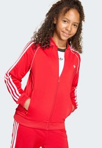 Κόκκινο τζάκετ με φερμουάρ, κατασκευασμένο από λείο ύφασμα με λευκές ρίγες στα μανίκια, διαθέτει ψηλό γιακά και πλαϊνές τσέπες.