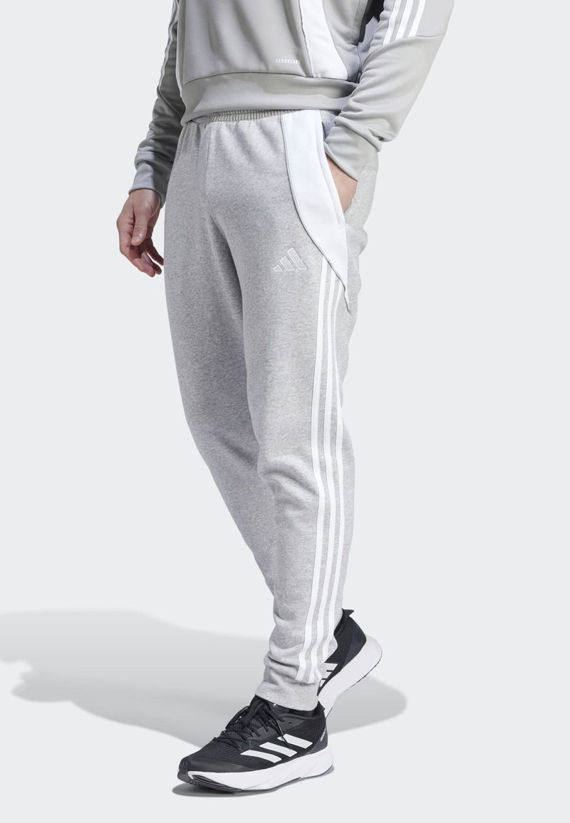 Pantalon de survêtement gris avec des rayures blanches sur les côtés et un logo Adidas, fabriqué en matériau doux. Associé à des chaussures de sport noires.