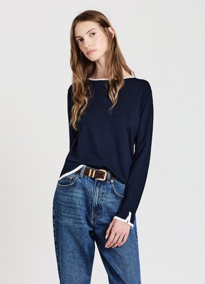 Navy long-sleeve knit top with white trim at the neckline and sleeve cuffs, paired with high-waisted blue jeans and a brown belt.